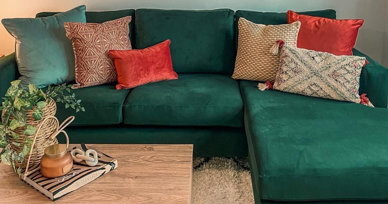 A green velvet sofa with layers of all different cushions in coral, natural and green colours. There are prints of oranges on the wall and a plant on the wooden coffee table. 