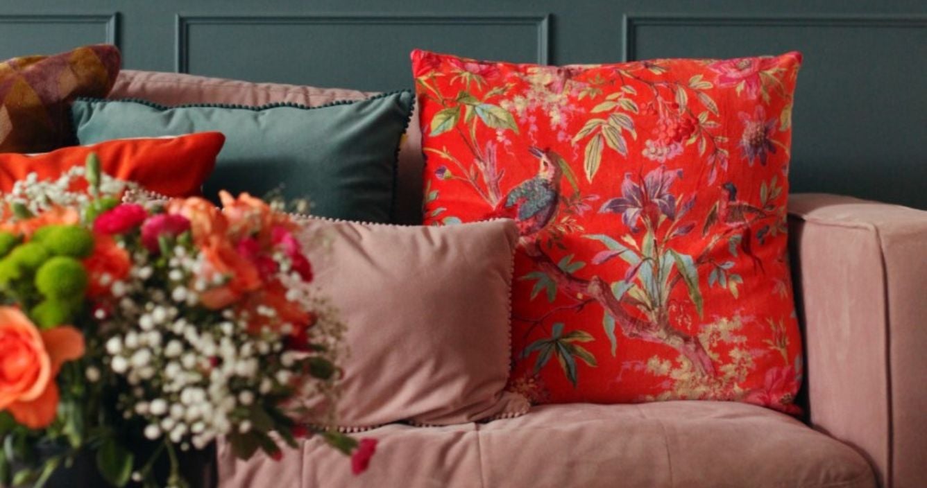 a pink velvet sofa with lots of velvet cushions on it, including a big red/orange cushion with a bird pattern on it. In the foreground there is a bunch of flowers.