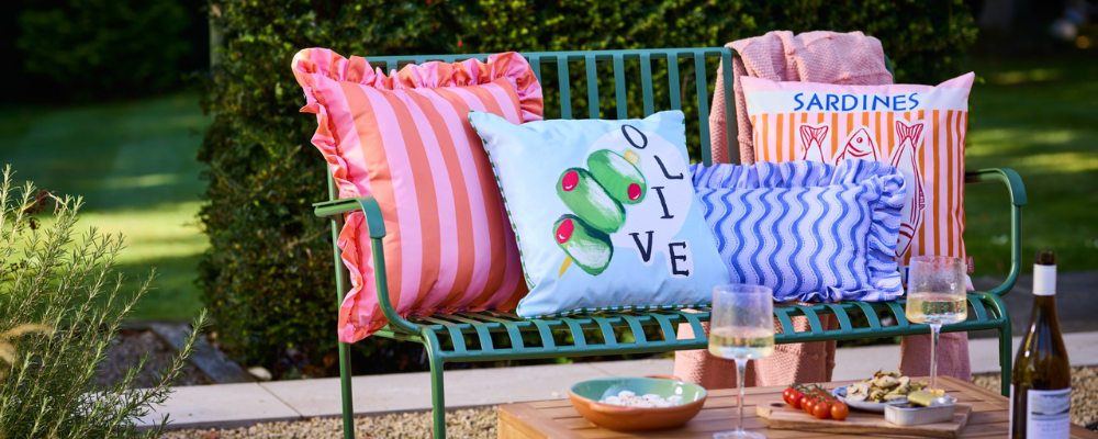 An outdoor bench with 4 outdoor cushions on with bright colourful prints including pink stripes, olives, purple squiggles and a sardine printed cushion