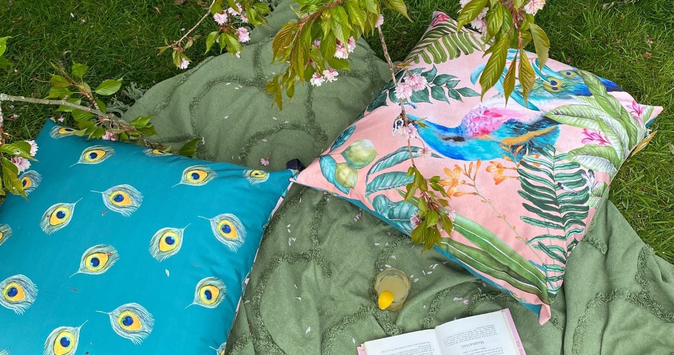 Two outdoor floor cushions with the same peacock design, laid on a green tufted throw in a garden, with both sides of the reversible design on display.