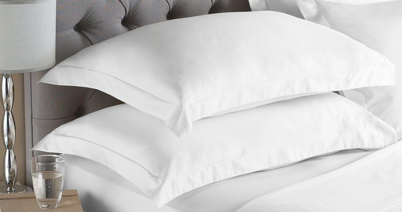 Four pillows with plain white cotton pillowcases arranged on a bed with white bedding and a grey button pleated headboard, next to a side table holding a table lamp and a glass of water.