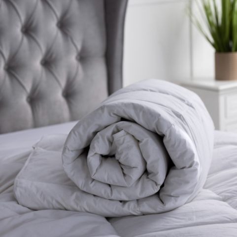 A luxury hotel quality duck feather quilt, rolled up and placed on top of a white mattress, with a grey pleated button headboard and a white side table holding a potted plant in the background.