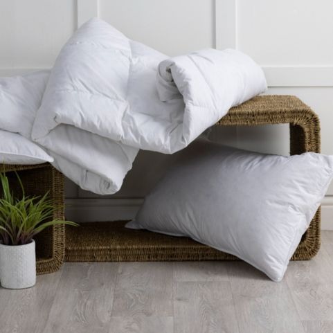 A white anti-allergy quilt and matching white pillow, scattered on woven wicker benches in front of a white wall and grey wood flooring, with a potted plant in the foreground. 
