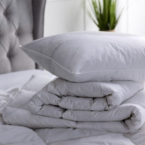 A luxury hotel quality duck feather pillow and matching quilt, arranged on a bed with a white mattress and a grey pleated button headboard, with a potted plant on a side table in the background.