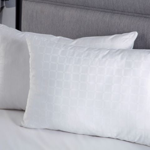 Two white, luxury hotel quality, polyester-filled pillows, stood up in front of a grey fabric headboard on a bed.
