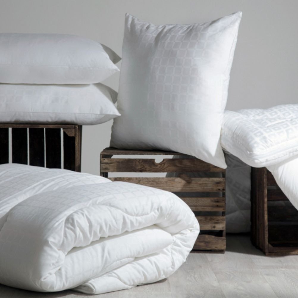 A collection of white, hotel quality bedding items, including two sleeping pillows, two quilts and a decorative cushion, arranged on wooden plank stands.