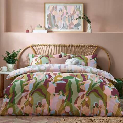 A multicoloured duvet cover set with a fluid abstract design of human forms, arranged on a bed with a woven headboard, in a room with blush pink walls and various decorative ornaments.