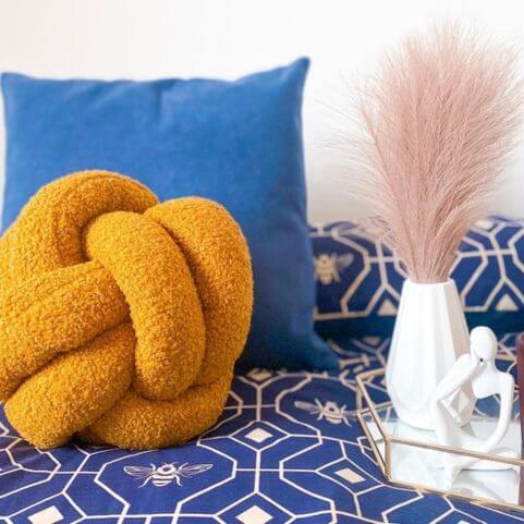 A yellow bouclé knot cushion placed in front of a plain blue bed cushion, next to a metal and glass tray holding an ornament on a geometric duvet cover set with a bee design.