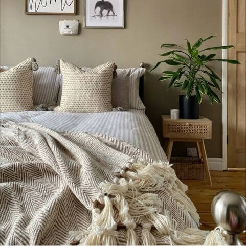 A tasseled bed throw with a woven herringbone pattern, placed messily over a striped duvet cover set with coordinating bed cushions, in a room with green walls and a side table holding an indoor plant.