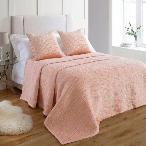 A luxury blush pink bedspread made on a bed with a white headboard, in a bedroom with white paneled walls, a white fur rug and a decorated side table.