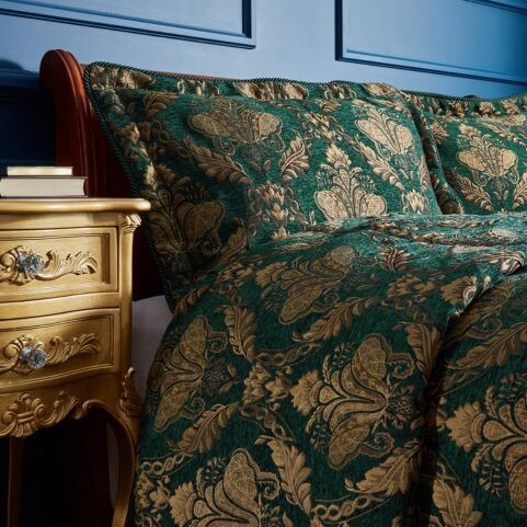 Two luxury green pillow shams with jacquard gold designs, placed on a bed with a wooden bedframe next to a gold side table, in a bedroom with blue paneled walls.