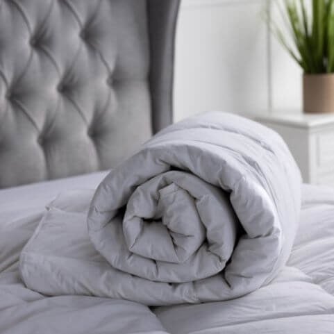 A luxury hotel quality white quilt with a duck feather filling, rolled up and displayed on a bed with a grey pleated bedframe.