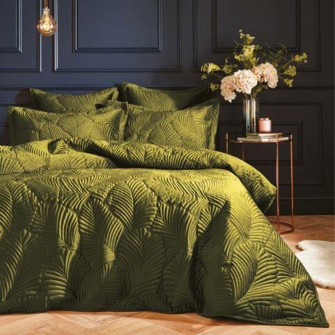A polyester duvet cover set with a leafy quilted velvet design in a moss green shade, made on a bed in a room with navy walls and a decorated side table.