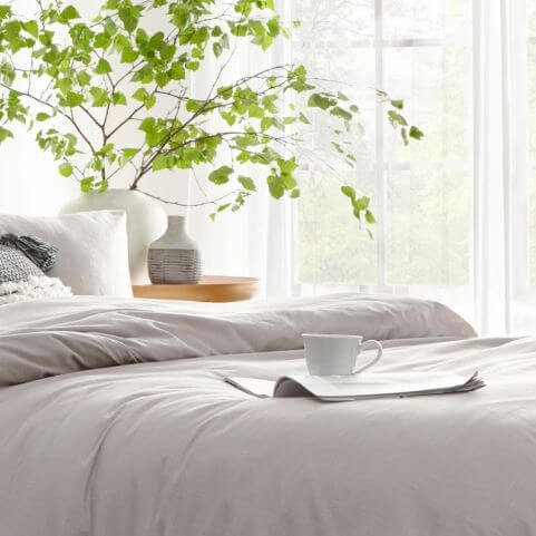 A linen look duvet cover set in a light grey shade, made on a bed in a room with a decorated side table, a large potted plant and a magazine holding a coffee cup.