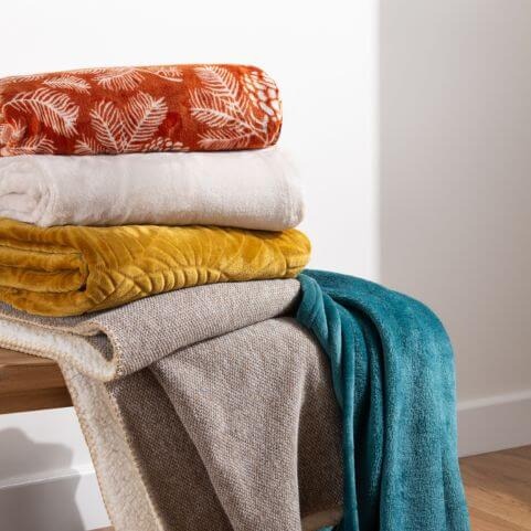 A selection of fleece and sherpa fleece throw blankets, folded and stacked on a wooden bench, and draping to the floor.