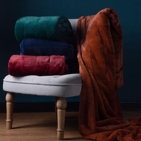 A collection of luxurious faux fur throw blankets, folded and displayed on an upholstered grey chair, with a rust coloured throw draped over the side.