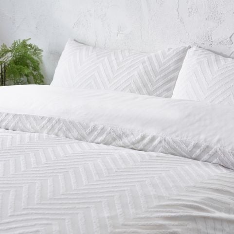 A white cotton duvet cover set with a tufted chevron design, made on a bed next to an indoor plant in front of a white background.