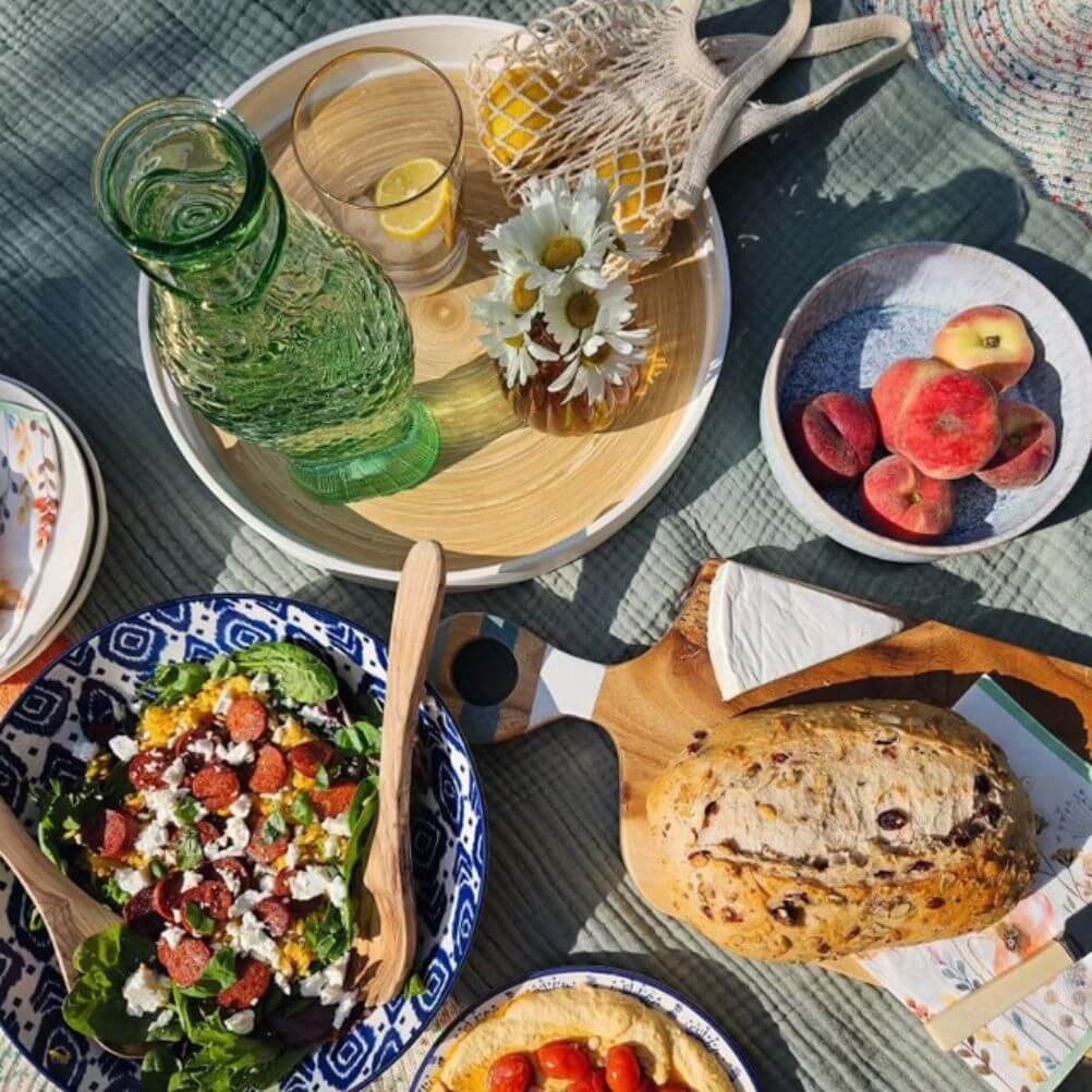 A picnic spread of seeded bread, Greek salad, fruit and water, laid on a crinkle cotton throw blanket outdoors.