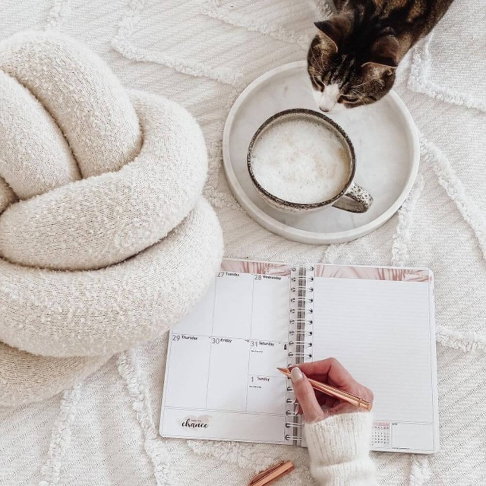 A person writing in a planning journal laid out on a white bed, surrounded by a white boucle knot cushion, a marble effect tray holding a cup of coffee, and a curious cat.