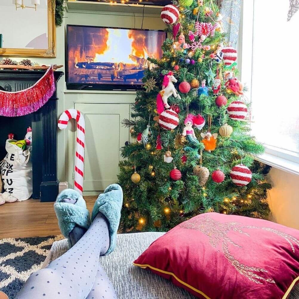 A person relaxing in a festively designed living room, including a red stag cushion, a decorated tree, a giant candy cane and a television displaying a Yule log fire.