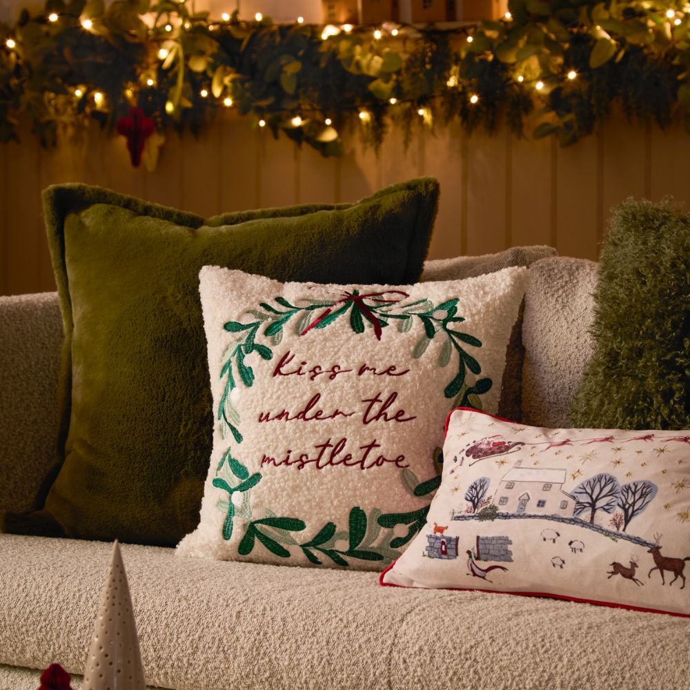 A Christmas set up with twinkling lights, a green faux fur cushion, a kiss me under the mistletoe cushion and a Cotswold scene rectangle cushion