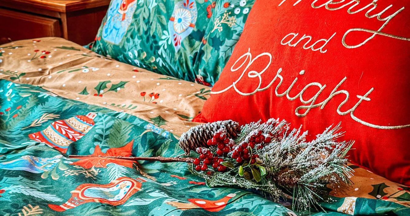 A green festive duvet cover set with a traditional design of baubles and trinkets, arranged on a bed with a red scatter cushion and some festive foliage.