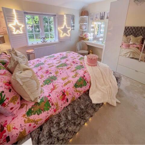 A bedroom with festive pink bedding featuring a design of leopards among Christmas trees, a white faux fur throw, neutral scatter cushions and novelty star-shaped lighting fixtures.