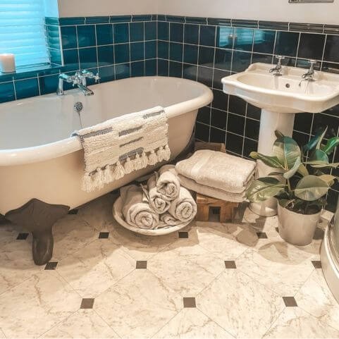 A bathroom with neutral and teal tiling, decorated with folded and rolled up towels and a neutral bath mat.