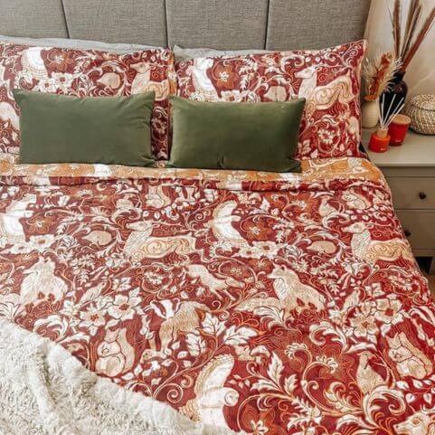 A red Christmas duvet cover set with a design of woodland animals and florals, presented on a bed with olive green scatter cushions and a neutral faux fur throw.
