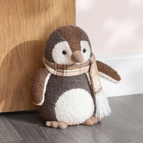 A novelty penguin door stop made with soft velvet and bouclé fabric, stood against a wooden door in front of a neutral background.