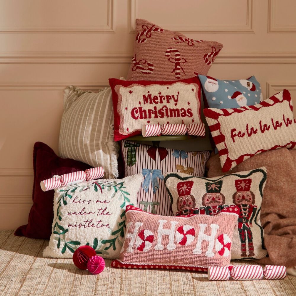 A stacked collection of festive cushions in pinks, blues, greens and reds. One says Merry Christmas, one says kiss me under the mistletoe, one says ho ho ho and one says fa la la la la.