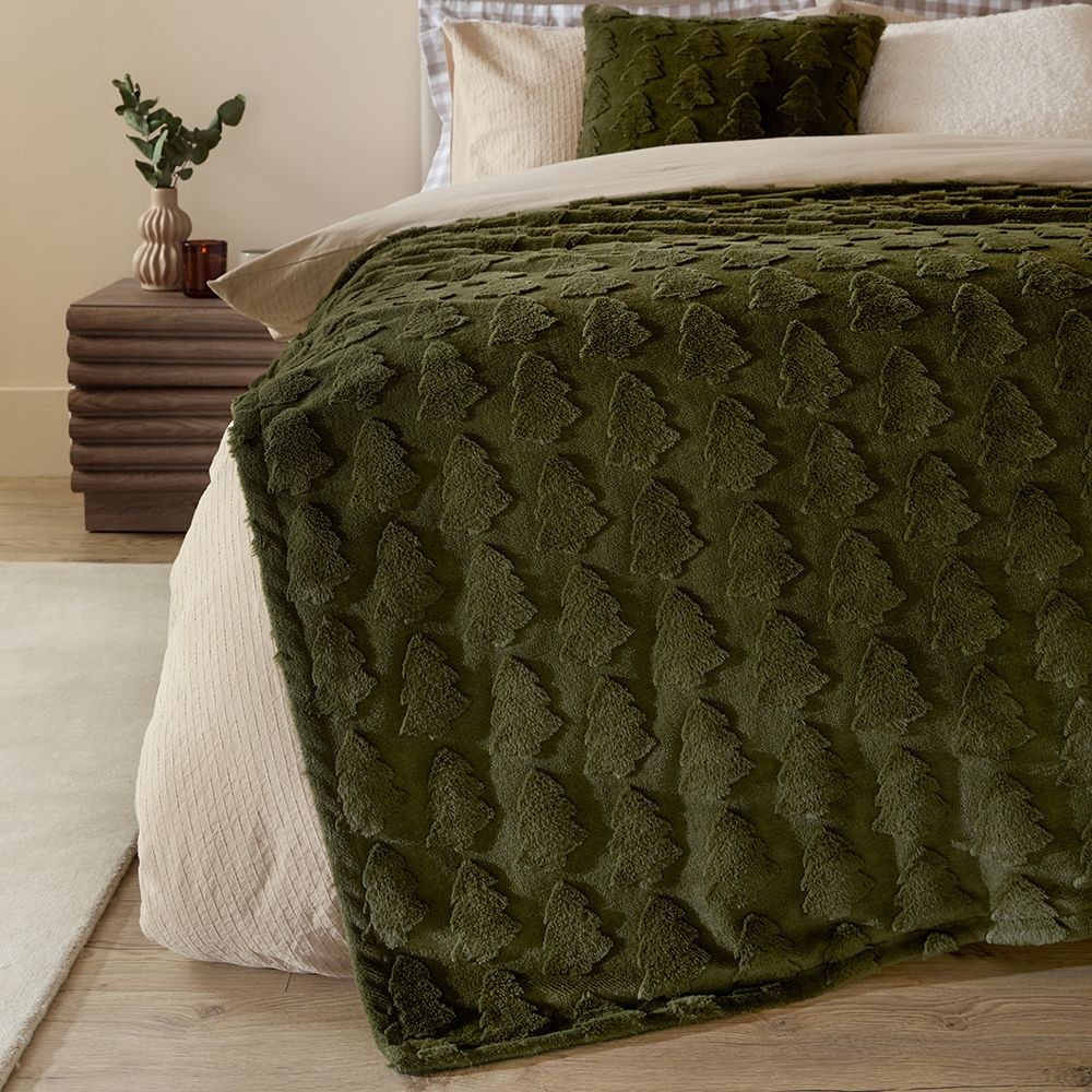 A bed with a white duvet cover on, with a green faux fur throw with tufted Christmas trees on it draped along the bottom