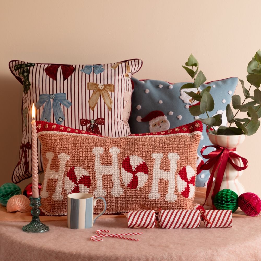 A Christmas table set up with three festive cushions. One is striped with bows on, one is blue with Santa faces, one is pink and rectangular, and says ho ho ho. There is a lit candle stick, a red and white Christmas cracker, candy canes, and a white and blue striped mug on the table.