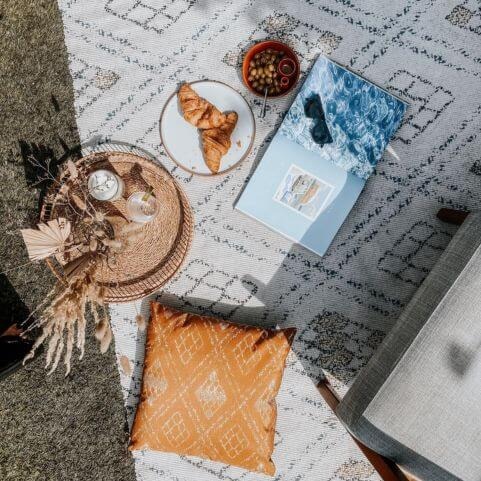 A boho style outdoor rug with a geometric diamond design, lying on a grass surface with a coordinating outdoor cushion, accessories and plates of food.