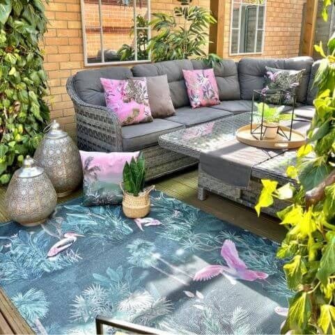 A polyester indoor/outdoor rug with a printed design of exotic birds and green forest florals, laid on a wooden decking surface along with an outdoor sofa, table and coordinating outdoor cushions.
