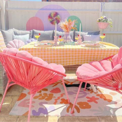 A polyester indoor/outdoor rug with an abstract multicoloured floral design, laid on a stone patio with two pink patio chairs and an outdoor sofa with coordinating outdoor cushions.