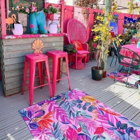 A polyester indoor/outdoor rug with a multicoloured design of vibrant tropical florals, laid on a wooden decking among a maximalist outdoor scene.