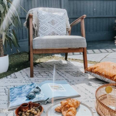 A boho style polyester indoor/outdoor rug with a geometric diamond design, laid on a grass surface with various summer items and a chair holding a matching outdoor cushion.