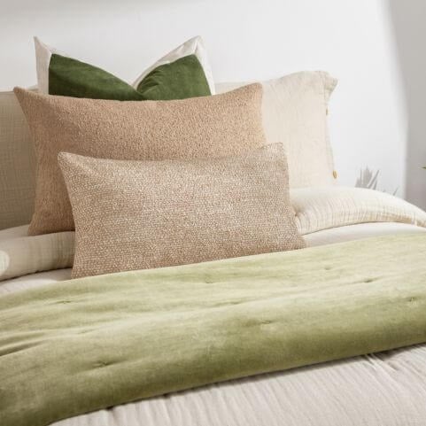 A minimalist biophilic bed decorated with a neutral muslin duvet cover, a moss cotton bedspread, brown bouclé cushions and an olive green velvet cushion.