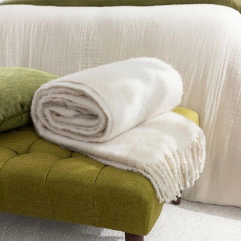 A faux mohair throw in a natural off-white shade, resting on an earthy green bedroom bench in a biophilic style bedroom.