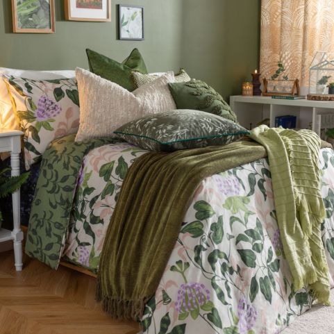 A maximalist biophilic bedroom decorated with a multicoloured floral duvet set, two green throws and a range of textured scatter cushions in green and white hues.
