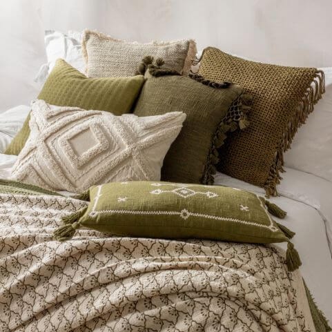 A selection of tufted, tasselled, fringed and jacquard scatter cushions in earthy green and natural shades, arranged on a white duvet set with two throws.