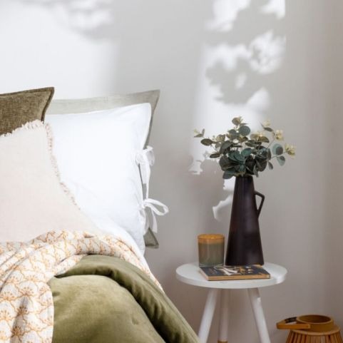two white cushions and olive faux fur throw next to a bedside table with plant and candle