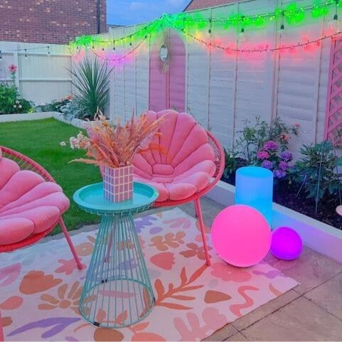 A garden scene featuring a floral outdoor rug laid on a stone patio, under two pink patio chairs, an outdoor table and various accessories.