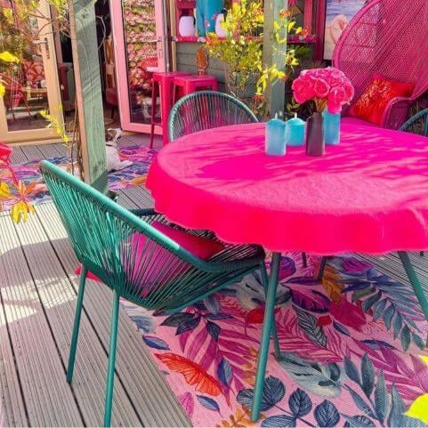 An outdoor scene of a wood deck decorated with a vibrant outdoor rug, green garden furniture and a hot pink table cloth.