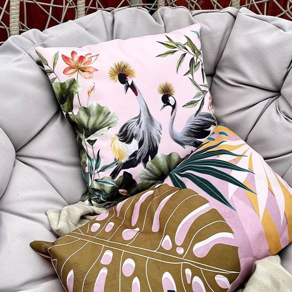 Two outdoor cushions with printed crane and leafy floral designs, arranged on a garden chair.