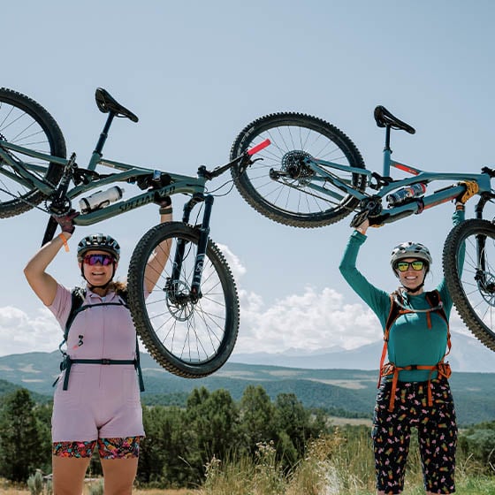 Two women holding their mountain bikes over their heads