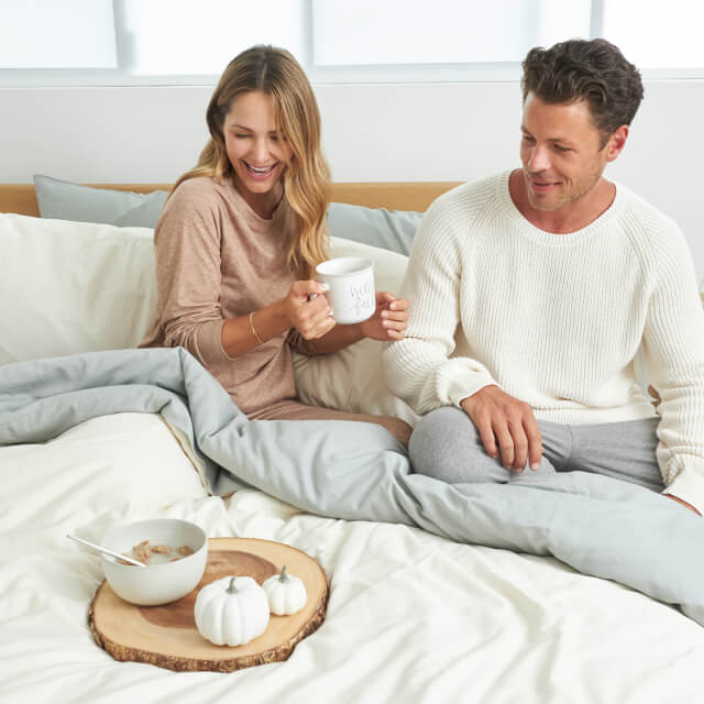 A man and woman sitting up in bed with the Slumber Cloud Down Comforter and drinking coffee