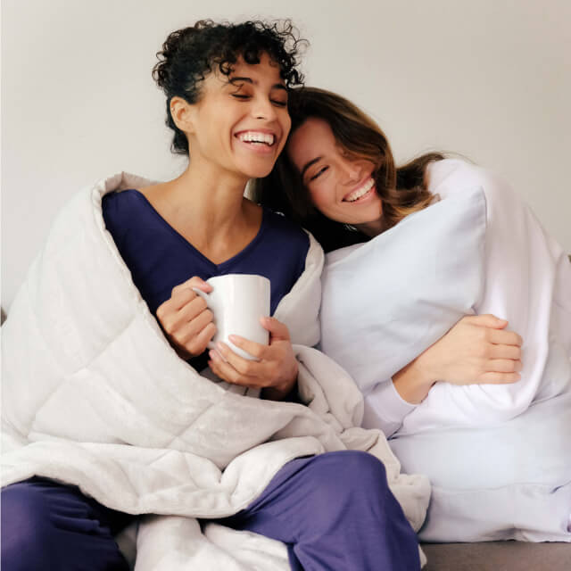 Two women smiling drinking coffee and wrapped in a Slumber Cloud Plush Throw in fog and clutching a pillow with the Performance Pillowcases