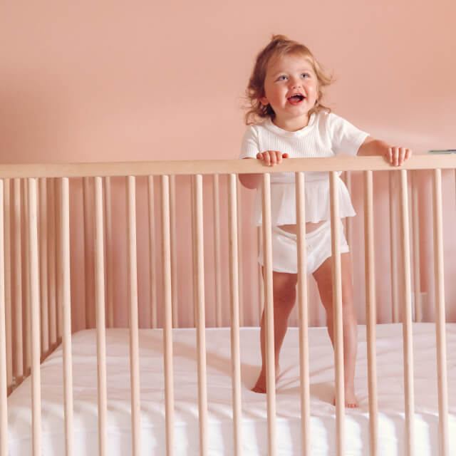 A young child smiling and standing in her crib with the Slumber Cloud Core Mattress Protector - Crib